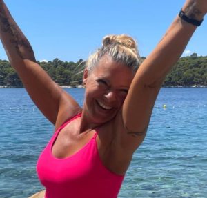 Claudia Hess fröhlich lächelnd vor einem See mit den Armen oben